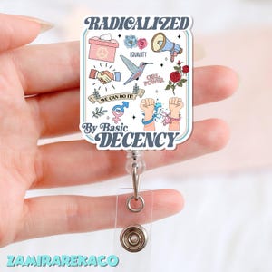 Może przedstawiać: Biała i jasnoniebieska smycz na identyfikator z napisami "RADICALIZED" i "By Basic DECENCY". Smycz na identyfikator zawiera ilustracje urny wyborczej, znaku pokoju, uścisku dłoni, kolibra i róż. Zawiera również tekst "WE CAN DO IT!".