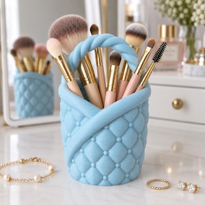 Peut inclure: Un porte-pinceaux de maquillage bleu clair avec une texture matelassée et une anse tressée. Le support contient divers pinceaux avec des viroles dorées et des poils beiges. Le récipient présente un design croisé sur le dessus.