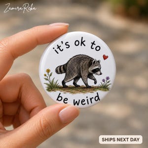 Op de afbeelding: Een ronde witte button met een centrale illustratie van een grijze en zwarte wasbeer die door gras met kleine bloemen loopt. De tekst "it's ok to be weird" staat in zwarte letters boven en onder de wasbeer met een klein rood hartje.