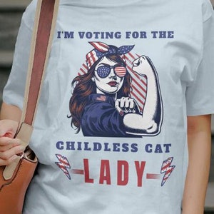 May include: Light blue t-shirt with a graphic of a woman wearing a red, white, and blue bandana and sunglasses. The text "I'm voting for the childless cat lady" is printed on the shirt.