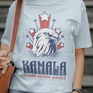 May include: Light blue t-shirt with a graphic of an eagle with stars and stripes. The text "Kamala Champion for Justice" is printed on the shirt.