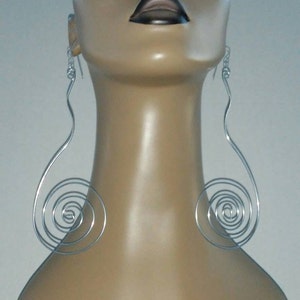 Op de afbeelding: Een mannequin met een paar zilveren spiraalvormige oorbellen. De oorbellen zijn lang en hangen van de oorlelletjes. De mannequin heeft een neutrale uitdrukking en kijkt recht vooruit.