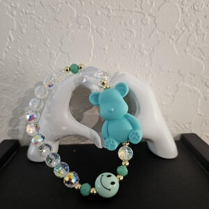 Puede incluir: Un brazalete de cuentas turquesa con un dije de oso de peluche turquesa y un dije de cara sonriente turquesa.