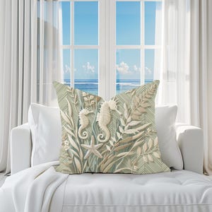 Pode incluir: Almofada decorativa com design de tema marinho. A almofada apresenta dois cavalos-marinhos, uma estrela-do-mar e elementos botânicos em tons de branco, bege e verde. Está colocada num sofá branco perto de uma janela.