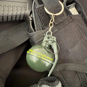 Può includere: Un portachiavi a granata verde con catena e anello dorati. Il portachiavi è attaccato a una borsa tattica nera. La granata ha un testo giallo che recita "GRADE HANDFRAGDELAYMQ" e numeri.