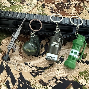 Könnte beinhalten: Eine Sammlung von Miniatur-Schlüsselanhängern im Militärstil. Die Schlüsselanhänger umfassen ein kleines Gewehr, eine grüne Granate, eine schwarze M18-Rauchgranate und eine grüne Granate mit weißen Akzenten. Präsentiert auf einem Tarnmuster-Hintergrund.