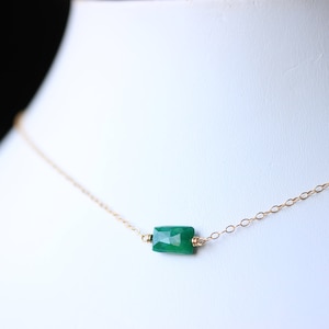 May include: A delicate gold chain necklace featuring a rectangular, emerald-green gemstone pendant. The gemstone is the focal point, suspended by small gold links. The necklace is displayed on a white surface.