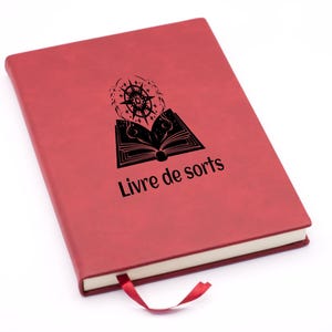 Peut inclure: Un journal relié en cuir rouge avec une illustration noire d'un livre ouvert et une étoile sur la couverture. Le texte "Livre de sorts" est imprimé sur la couverture.