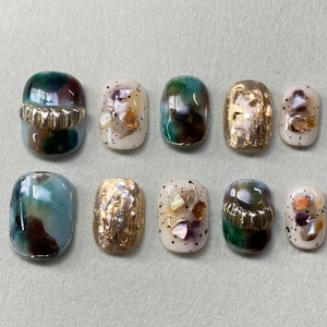 Puede incluir: Diez diseños de uñas diferentes que presentan una variedad de colores y patrones, incluyendo verde, marrón, dorado y blanco. Los diseños incluyen patrones abstractos, conchas marinas y purpurina.