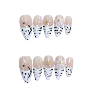 Puede incluir: Un conjunto de uñas postizas en forma de almendra. Las uñas presentan una base nude con puntas blancas y diseños negros. Algunas uñas tienen una estrella dorada o un acento de flor rosa. Las uñas están diseñadas para una manicura de moda.