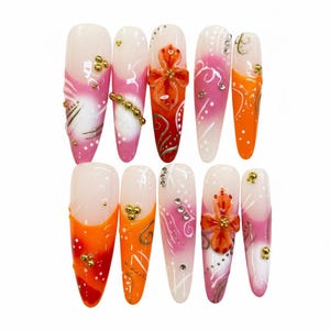 Puede incluir: Un conjunto de diez uñas artificiales con diversos diseños. Las uñas presentan una combinación de colores, incluyendo blanco, rosa y naranja, con adornos dorados y plateados. Algunas uñas tienen acentos florales.
