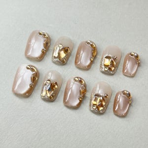 May include: Ten beige false nails with gold and clear rhinestones.