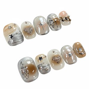 Puede incluir: Un conjunto de diez uñas artificiales con diversos diseños. Las uñas presentan una mezcla de colores plateado, marrón y blanco, con detalles de estrellas y corazones. Algunas uñas tienen un efecto marmolado, otras adornos metálicos.