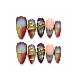 Puede incluir: Un conjunto de diez uñas postizas en forma de almendra. Las uñas presentan un diseño degradado con colores como el rojo, el amarillo y el azul. Algunas uñas tienen estrellas doradas, adornos dorados y detalles de perlas. Las uñas están dispuestas sobre una superficie blanca.
