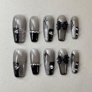 Puede incluir: Conjunto de diez uñas postizas grises y negras con purpurina plateada y detalles negros. Algunas uñas tienen un lazo negro, otras una estrella negra y otras una línea negra.