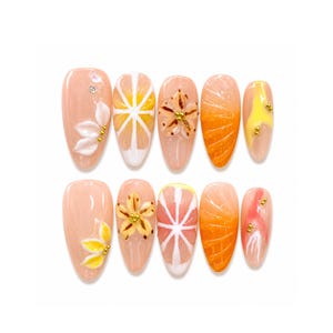 Puede incluir: Un conjunto de diez uñas postizas en forma de almendra. Las uñas presentan varios diseños, incluyendo flores, rodajas de cítricos y segmentos de naranja texturizados. La paleta de colores incluye tonos melocotón, naranja, amarillo y blanco, con detalles dorados.