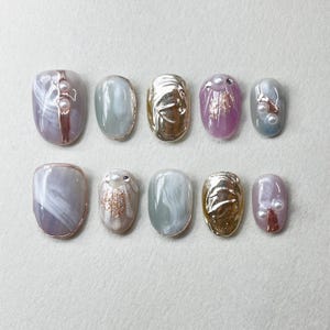 May include: Ten oval-shaped false nails with different designs. Some nails have a white and grey swirl pattern, others have gold leaf, and some have pearls and glitter.