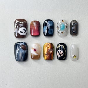 May include: Ten different nail designs featuring various colours, patterns, and textures. The designs include a bear face, a red and gold ombre, a blue and white swirl, a white design with blue and green stones, a brown design with a black swirl, a black and white swirl, a white design with a red stone, a gold design with brown stones, a black design with a white cross, and a white design with a gold stone.