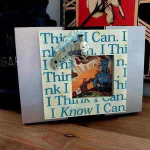 May include: A framed print with the text "I Think I Can. I Know I Can." features a colorful illustration of a train. A small train figurine is attached to the top of the frame. The frame is light gray.