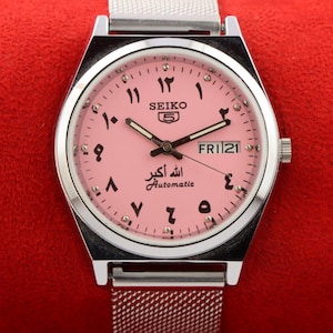 Może przedstawiać: Automatyczny zegarek Seiko 5 z różową tarczą i arabskimi cyframi. Zegarek ma srebrną kopertę i bransoletę z siatki. Dzień i data są wyświetlane na godzinie 3. Zegarek jest na czerwonym tle.