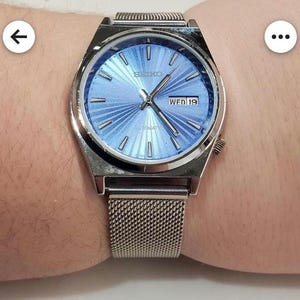 May include: A silver wristwatch with a light blue face and a mesh strap. The watch face shows the day and date, with the word "SEIKO" at the top. The watch hands and hour markers are silver. The watch is on a wrist.
