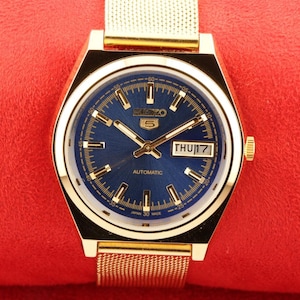 May include: A gold-tone Seiko automatic wristwatch with a blue face and a gold metal band. The face has gold-tone hour markers and hands, a date window, and the text "SEIKO AUTOMATIC" and "JAPAN 30 MADE".