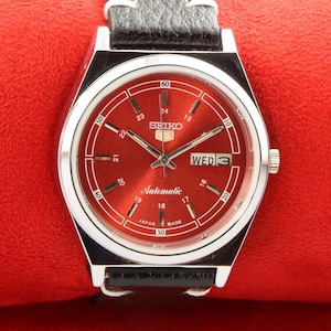 Peut inclure: Une montre-bracelet argentée avec un cadran rouge et un bracelet en cuir noir. Le cadran de la montre comporte des marques blanches et le texte "SEIKO Automatic Japan Made" imprimé dessus. Le jour de la semaine est affiché comme "WED 3".