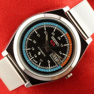 May include: A silver-toned Seiko automatic wristwatch with a black face, featuring a day-date display and a mesh band. The watch face has blue and orange accents, with the text "SEIKO" and "AUTOMATIC". The watch is set against a red background.