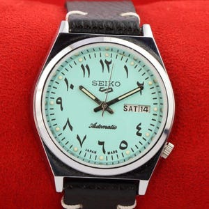Könnte beinhalten: Eine silberfarbene Armbanduhr mit einem hellblauen Zifferblatt und schwarzen arabischen Ziffern. Das Zifferblatt der Uhr enthält auch die Wörter "SEIKO Automatic" und "SAT 14".