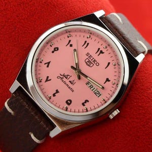 May include: A Seiko automatic wristwatch with a pink face and Arabic numerals. The watch has a silver case and a brown leather strap. The face also displays the day and date, and the text "الله أكبر Automatic".