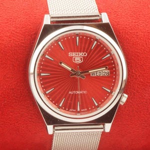 May include: A Seiko 5 automatic watch with a red dial and silver-tone case and mesh band. The dial features a sunburst pattern, day and date display, and the text "SEIKO 5". The watch is displayed on a red background.