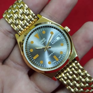 Op de afbeelding: Een goudkleurig Seiko automatisch polshorloge met een zilveren wijzerplaat en gouden wijzers. De wijzerplaat toont de dag van de week en de tekst "Diver's 200M". Het horloge heeft een goudkleurige band.
