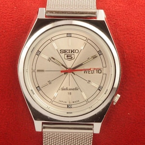 Op de afbeelding: Zilverkleurig Seiko 5 automatisch horloge met een lichtgekleurde wijzerplaat. Het horloge heeft een zilveren mesh band, een dag-datumweergave en de tekst "SEIKO 5 Automatic 18 JAPAN MADE". De wijzers van het horloge zijn zwart en rood.