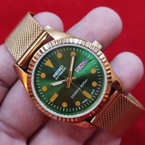 May include: Gold-toned Seiko automatic watch with a green dial and a mesh band. The watch face displays the day and date, with the text "SEIKO AUTOMATIC" and "DIVER'S 200m". The watch has a gold bezel and a gold-colored mesh band.