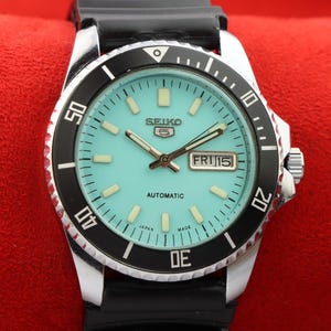 May include: A black and silver Seiko automatic wristwatch with a blue face and a black rubber strap. The watch face has white markings and the text "SEIKO 5 AUTOMATIC JAPAN MADE" and "FRI 15".