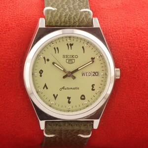 May include: A Seiko 5 automatic watch with a silver case and a light green face featuring Arabic numerals. The watch has a green leather band and displays "WED 20".