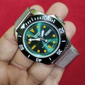 Pode incluir: Um relógio de mergulho automático Seiko em aço inoxidável com mostrador verde e moldura giratória preta. O relógio apresenta uma pulseira de malha e exibe o dia e a data. O mostrador diz "DIVER'S 200m" e "JAPAN".