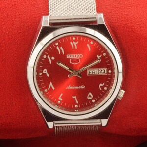 May include: A silver wristwatch with a red face and Arabic numerals. The watch has a silver metal band and says "SEIKO Automatic" and "FRI 23" on the face.