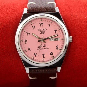 May include: A pink-faced Seiko 5 automatic wristwatch with Arabic numerals and a day of the week display. The watch face also features the text "Allah Akbar" and "Automatic".