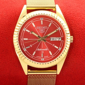 Peut inclure: Une montre-bracelet dorée avec un cadran rouge et un bracelet en métal doré. Le cadran de la montre comporte une fenêtre de date et le texte "SEIKO 5 Automatic 18 JAPAN MADE" imprimé dessus.