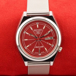 Puede incluir: Un reloj de pulsera automático Seiko 5 plateado con una esfera roja y una correa de metal plateado. La esfera del reloj tiene marcas blancas y una ventana de día y fecha.