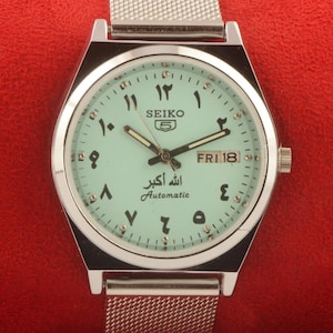 May include: A silver Seiko 5 automatic wristwatch with a light green face and Arabic numerals. The watch has a day and date display and a mesh strap. The watch face includes the text "SEIKO 5", "Automatic", and Arabic script.