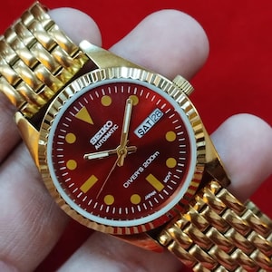 Puede incluir: Reloj automático Seiko dorado con esfera roja y detalles dorados. El reloj presenta una pantalla de fecha y una correa dorada texturizada. La esfera indica "SEIKO AUTOMATIC DIVER'S 200m".