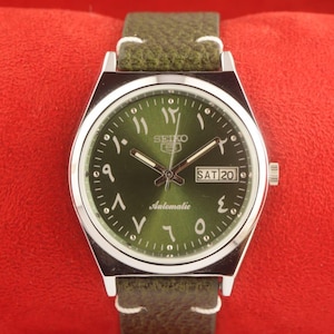 May include: A vintage Seiko 5 automatic watch with a green dial and Arabic numerals. The watch has a silver-tone case and a green leather strap. The day and date are displayed at the 3 o'clock position. The watch is set against a red background.