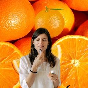 May include: A woman smelling a small bottle of essential oil, surrounded by fresh oranges and orange slices. The background is filled with whole oranges. The woman is wearing a white shirt and a black bracelet. A logo is visible in the bottom right corner.