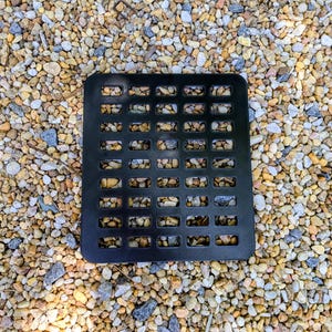 May include: A square black plastic drainage grate featuring a grid of rectangular cutouts. The grate has a smooth, flat surface with rounded corners and is positioned directly over a bed of small, multi-colored tan, gray, and white pebbles.