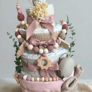 K&ouml;nnte beinhalten: Mehrst&ouml;ckige Windeltorte in Rosa, Creme und Braun, verziert mit B&auml;ndern, Holzperlen und einem geh&auml;kelten Fuchs. Enth&auml;lt einen Schnuller, Holzformen und Babyschuhe. Ein bezauberndes Geschenk f&uuml;r eine Babyparty.