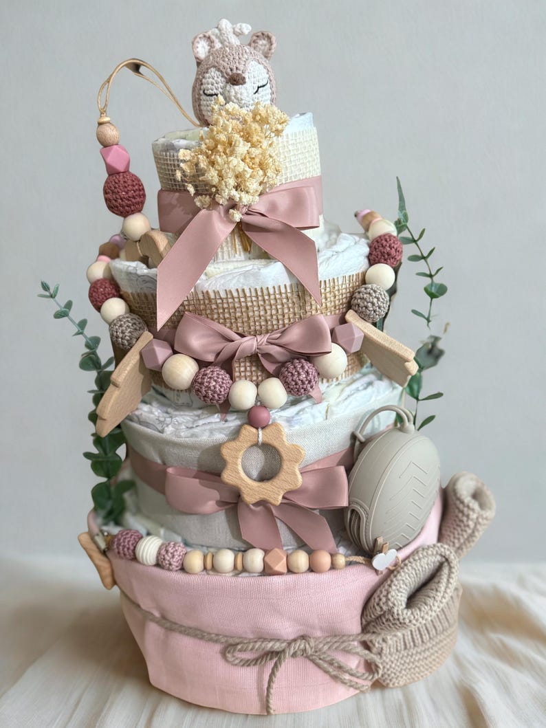 K&ouml;nnte beinhalten: Mehrst&ouml;ckige Windeltorte in Rosa, Creme und Braun, verziert mit B&auml;ndern, Holzperlen und einem geh&auml;kelten Fuchs. Enth&auml;lt einen Schnuller, Holzformen und Babyschuhe. Ein bezauberndes Geschenk f&uuml;r eine Babyparty.