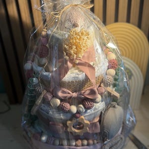 K&ouml;nnte beinhalten: Ein Geschenkpaket f&uuml;r Babys, verpackt in durchsichtigem Zellophan und mit einer rosa Schleife gebunden. Der Korb hat ein mehrst&ouml;ckiges Design und enth&auml;lt Babyartikel in Rosa-, Wei&szlig;- und Graut&ouml;nen. Enth&auml;lt einen Bei&szlig;ring aus Holz und dekorative Perlen.