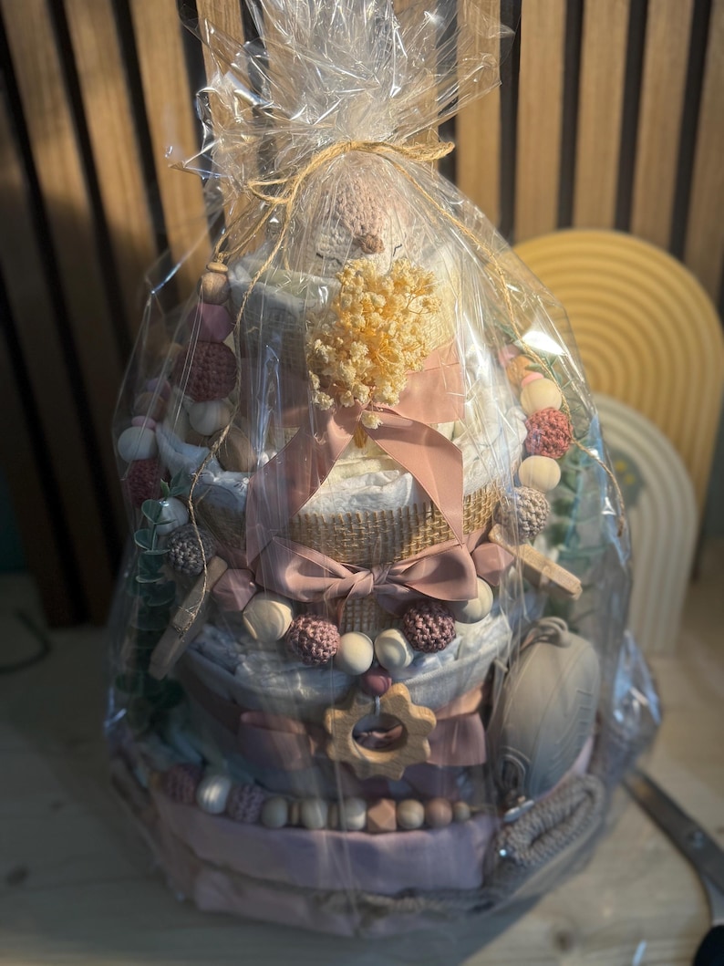K&ouml;nnte beinhalten: Ein Geschenkpaket f&uuml;r Babys, verpackt in durchsichtigem Zellophan und mit einer rosa Schleife gebunden. Der Korb hat ein mehrst&ouml;ckiges Design und enth&auml;lt Babyartikel in Rosa-, Wei&szlig;- und Graut&ouml;nen. Enth&auml;lt einen Bei&szlig;ring aus Holz und dekorative Perlen.